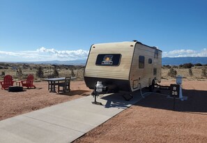 A recreational vehicle (RV) with a 'Observer Ranch' logo, parked on a dirt ground with a paved walkway, red Adirondack chairs, and a table. - Venus Camper Suite - Planet-themed camper suite with awning deck, sleeps up to three (Penrose)