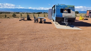 En blå husbil med texten ”Observer Ranch” på sidan står parkerad på en grusplan, och i närheten finns ett picknickbord och några stolar.