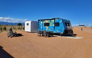 A blue and white camper parked on a gravel surface with a table and chairs nearby. - Earth Camper Suite - Planet-themed camper suite with awning deck, sleeps three (Penrose)