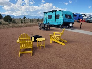 En campingplads med to gule Adirondack-stole, et lille rundt bord og en blå autocamper parkeret på grus.
