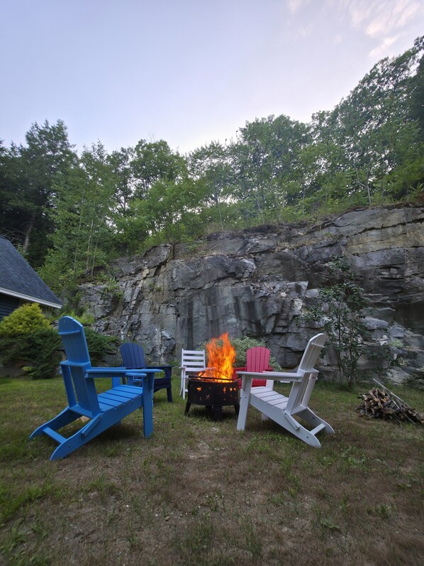 Patio trasero con fogón rodeado de sillas Adirondack, una silla blanca y un muro de piedra.