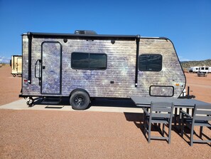 A stationary RV with a side door, a small window, and a table and chair set outside. - Mercury Camper Suite - Planet-themed camper suite with awning deck, sleeps three (Penrose)