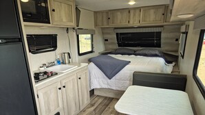 Interior de una caravana compacta con kitchenette, una cama con una manta azul y zona de comedor.