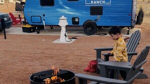 Una caravana azul con la marca 'OBSERVER RANCH' estacionada en un camping con mobiliario exterior y una fogata.