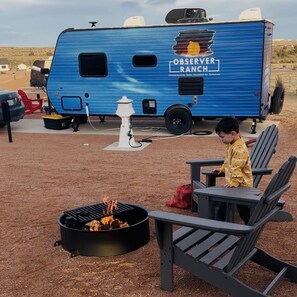 En blå autocamper med OBSERVER RANCH branding parkeret på en campingplads med udendørs siddepladser og bålsted.