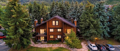 Un chalet à deux étages, doté de fondations en pierre, entouré d’une forêt dense, et comprenant un espace de stationnement pouvant accueillir plusieurs véhicules.