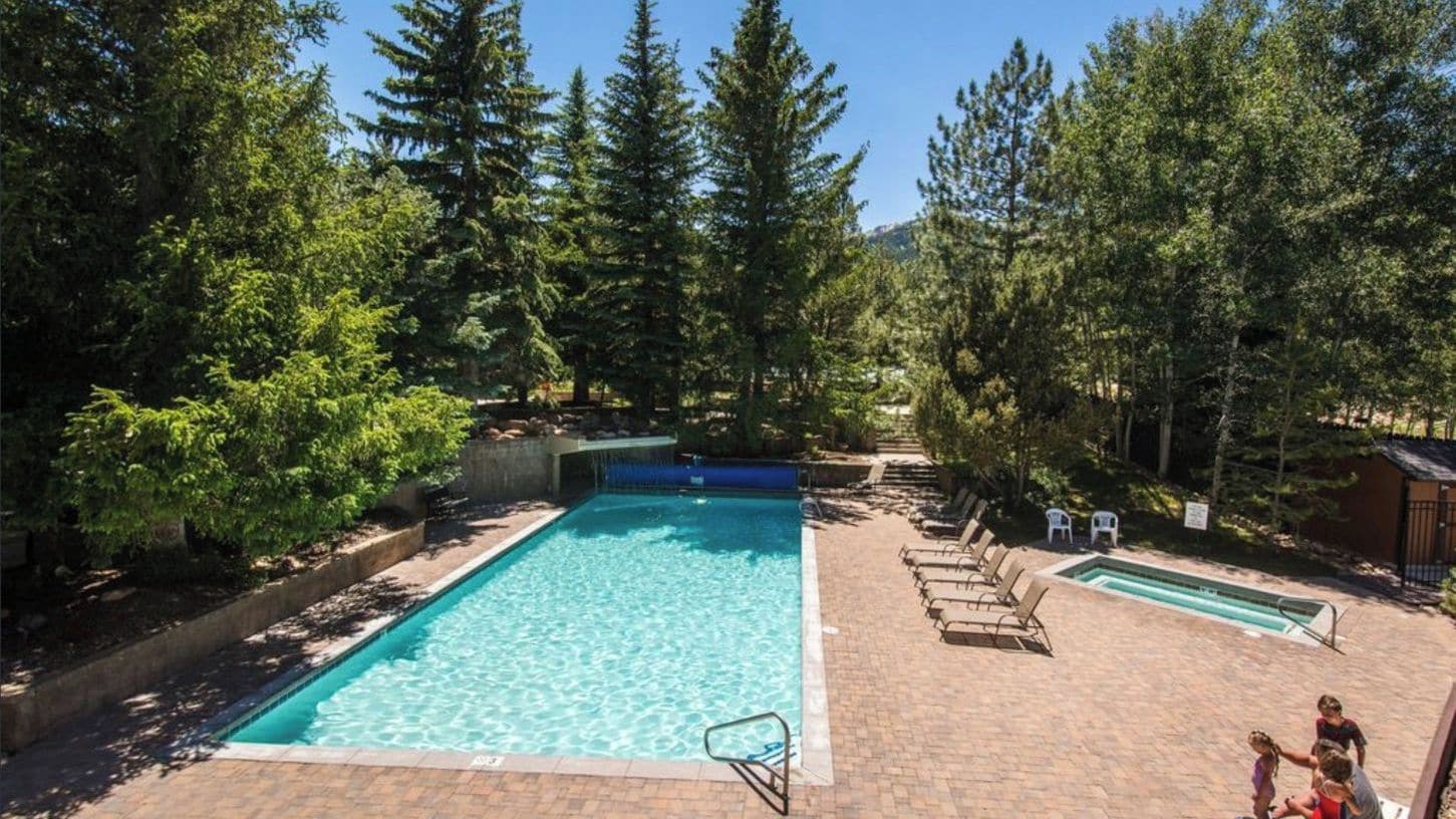 A rectangular swimming pool with a diving board, surrounded by trees and a brick patio.