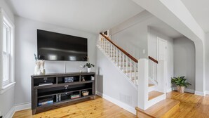 Een moderne woonkamer met een houten vloer, een flatscreen televisie aan de wand, een donkere houten entertainmentkast en een trap met witte leuningen.