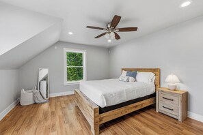 Un dormitorio con techo inclinado, ventilador de techo, cama con cabecera de madera, mesita de noche con lámpara y ventana con vista a follaje.