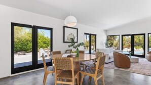 Ein modernes Interieur mit einem runden Esstisch, Holzstühlen, einer Couch und einem großen Fenster mit Blick auf Grünflächen.