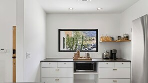A modern kitchen with white cabinetry, a built-in microwave, and a window with a framed picture of a desert landscape. - Casa del Cielo by Avantstay Private Pool + Spa + Desert Patio + Grill Mountain Views + Loungers (Palm Springs)