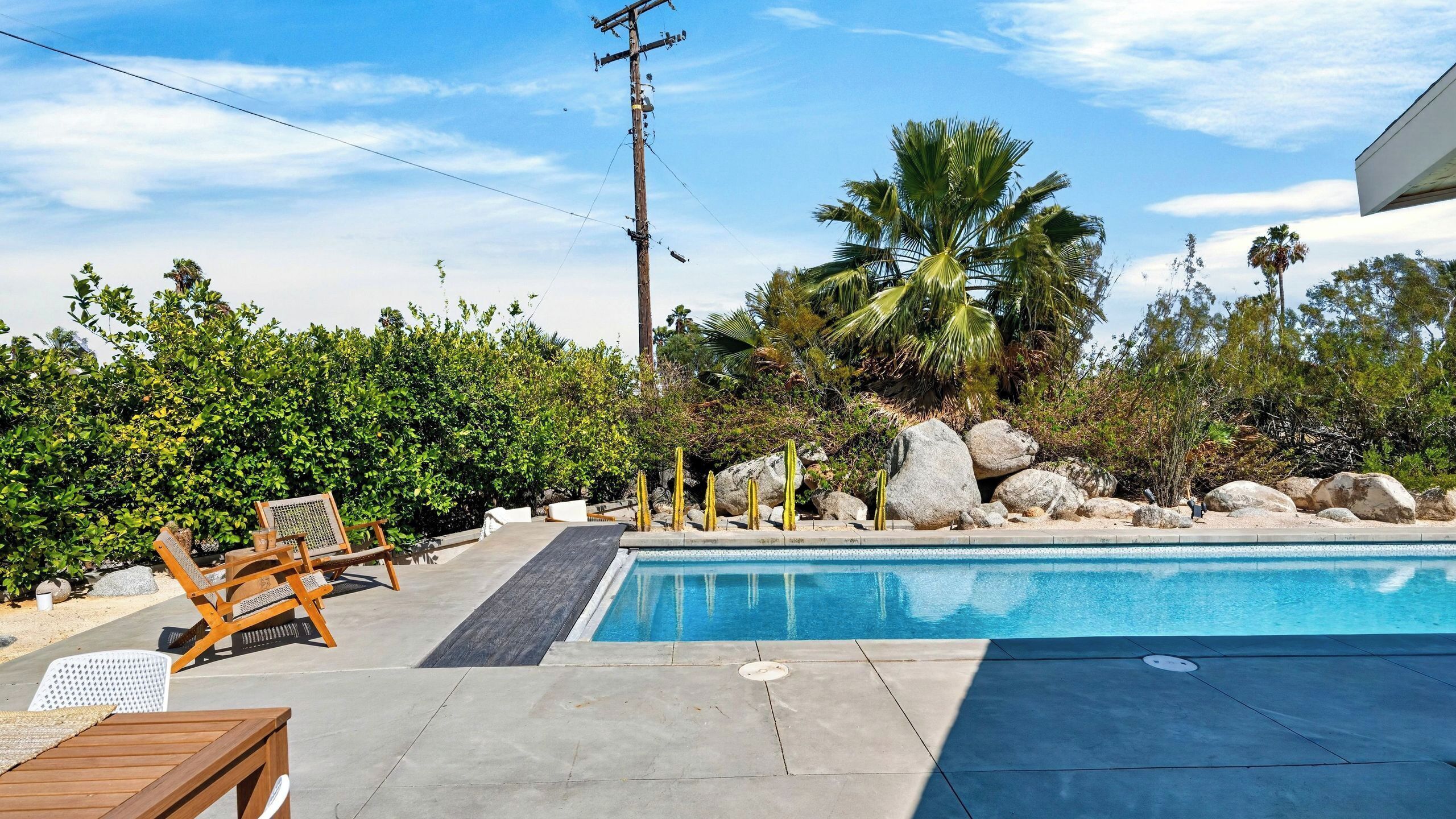 A swimming pool with a wooden deck, lounge chairs, and a rocky landscape with cacti and palm trees.