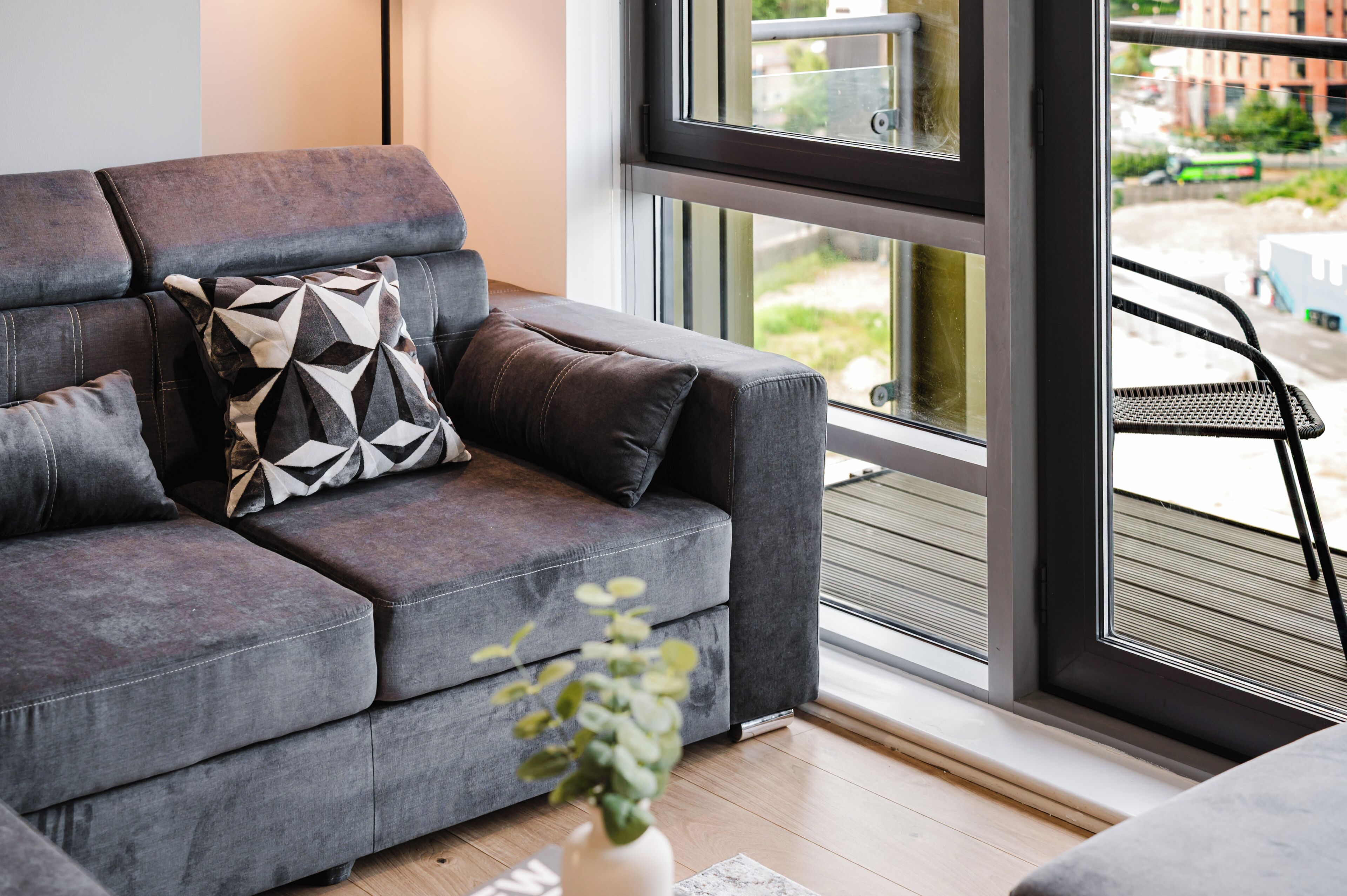 A grey sofa with a geometric pillow, a small plant, and a view of a cityscape through a window.
