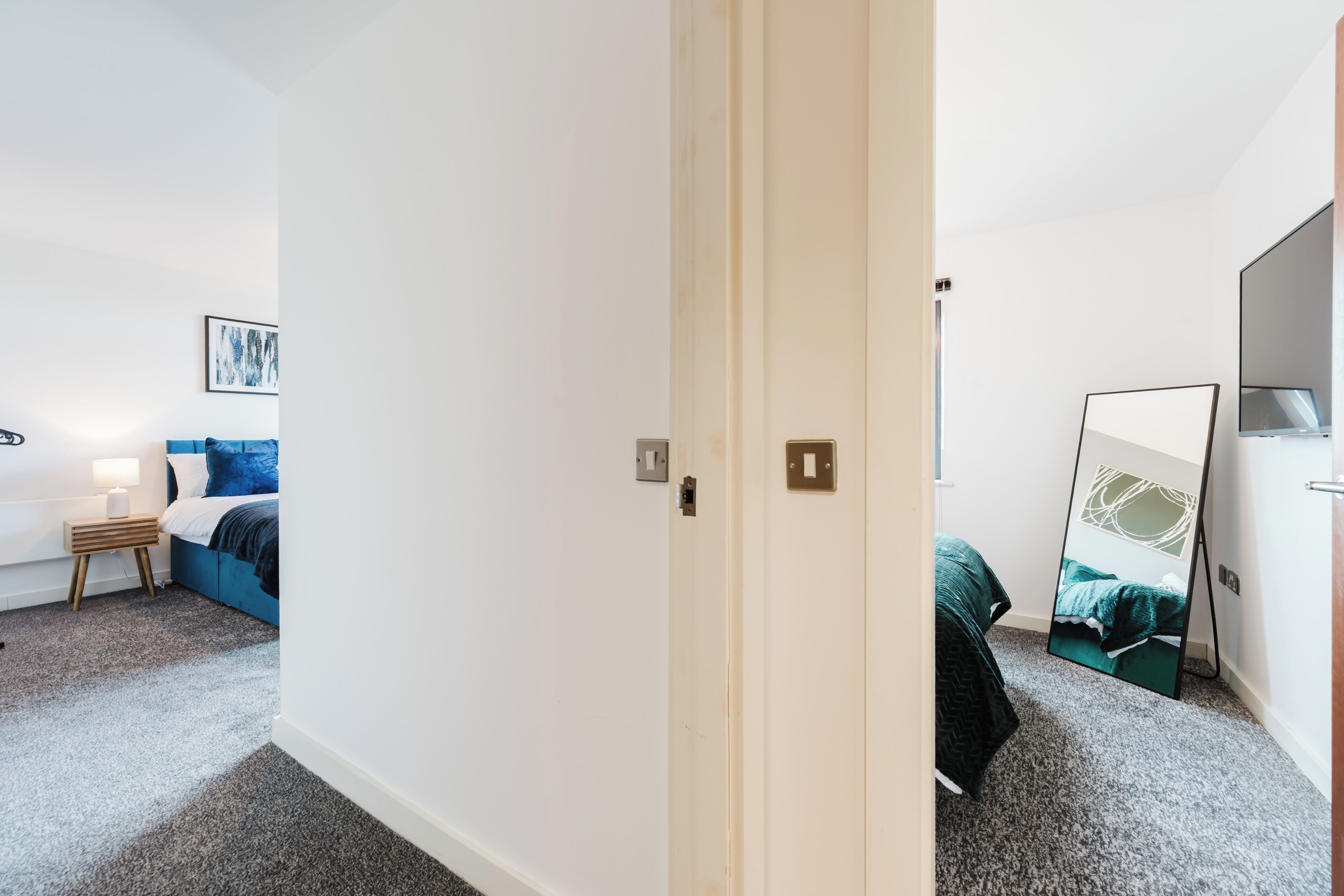 A hotel room with a bed, a bedside table with a lamp, a wall-mounted TV, and a door leading to another room.