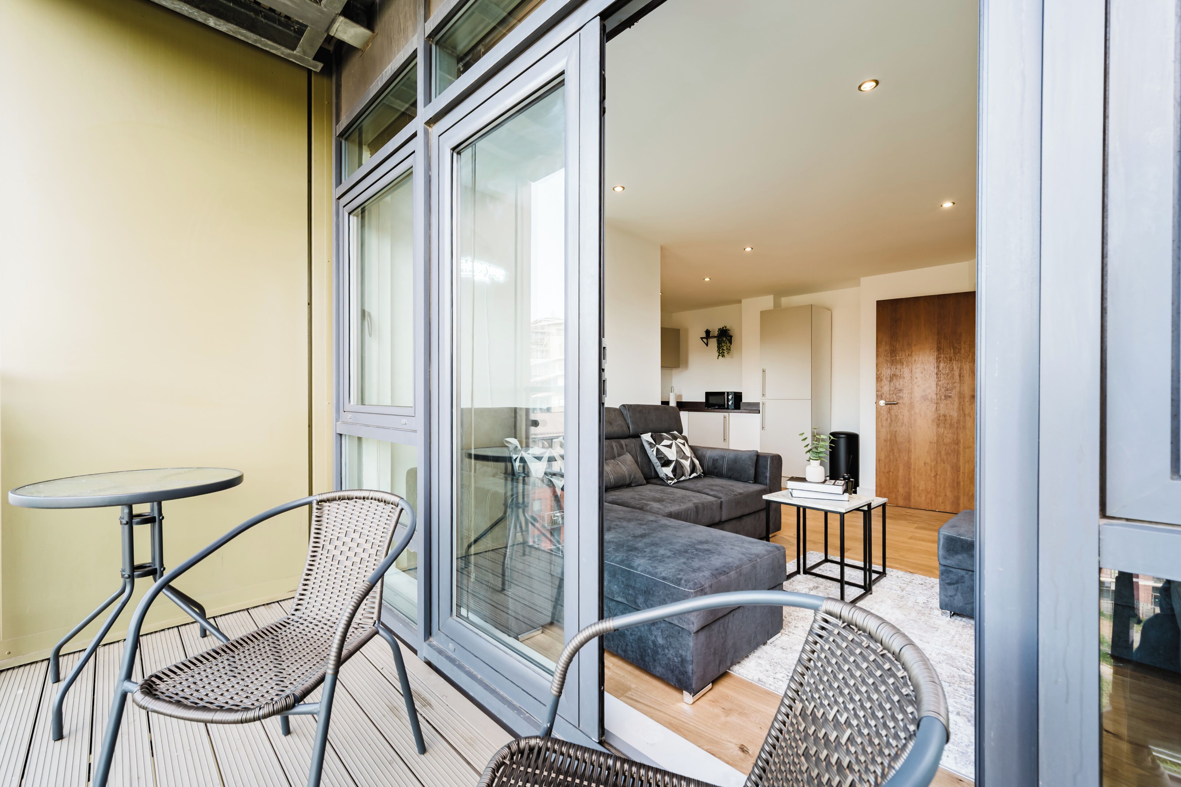 A balcony with two chairs and a table, a sliding glass door leading to a living area with a sofa, a coffee table, and a kitchen area visible through the door.