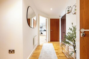 Un pasillo con suelo de madera, pared blanca, espejo redondo y puerta que da a una cocina.