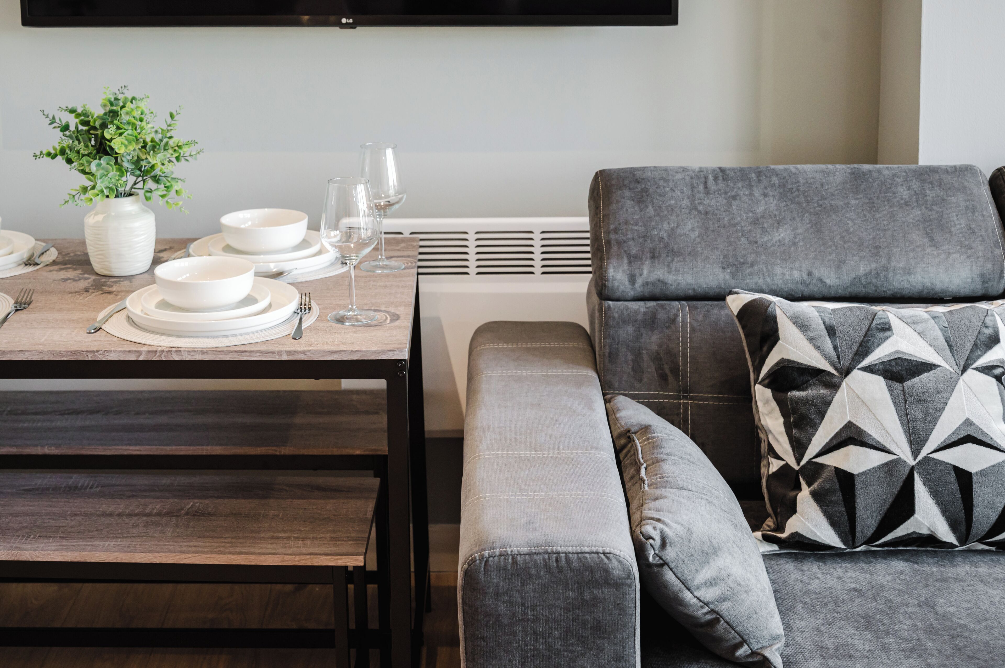 A dining area with a table set for four, a grey sofa with a patterned pillow, and a white vase with green plants.