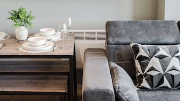 A dining area with a table set for four, a grey sofa with a patterned pillow, and a white vase with green plants.
