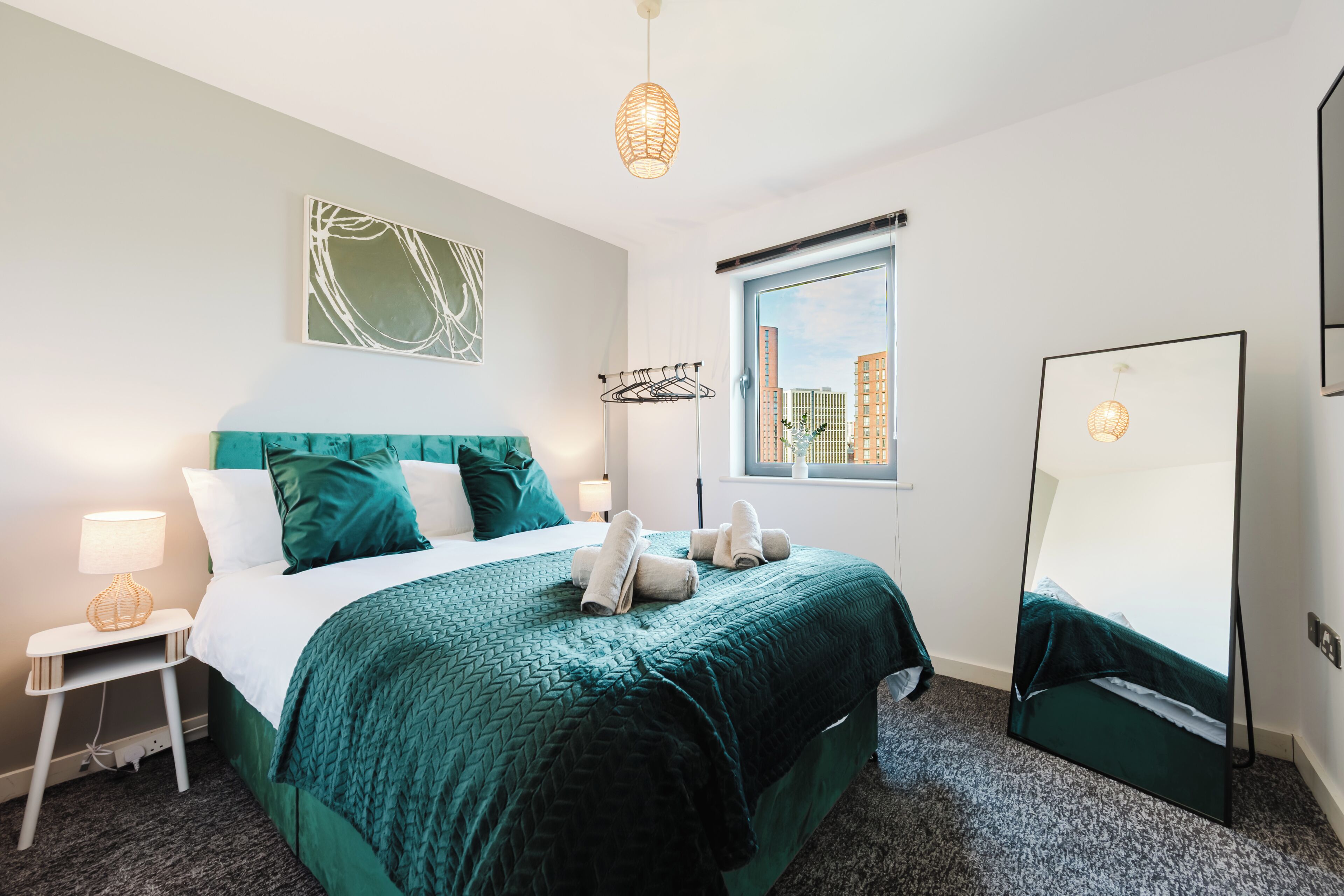 A hotel room with a bed, a window with a view of buildings, a bedside table with a lamp, and a mirror.