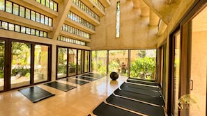 Amplo espaço interno com vários tapetes de yoga dispostos em fileiras, grandes janelas e vista para o verde lá fora.