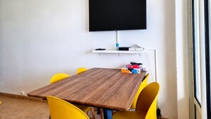 Uma sala de conferências com uma mesa de madeira, cadeiras amarelas, uma TV de tela plana montada e uma estante branca com dispositivos eletrônicos.