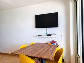 A conference room with a wooden table, yellow chairs, a mounted flat-screen TV, and a white shelf with electronic devices. - Le Mosala 5 - Climatisation - Vue mer (Saint-Leu)