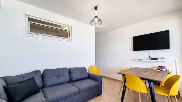 Uma sala de estar com sofá, uma mesa de jantar com cadeiras, uma televisão de tela plana fixada na parede e um lustre.