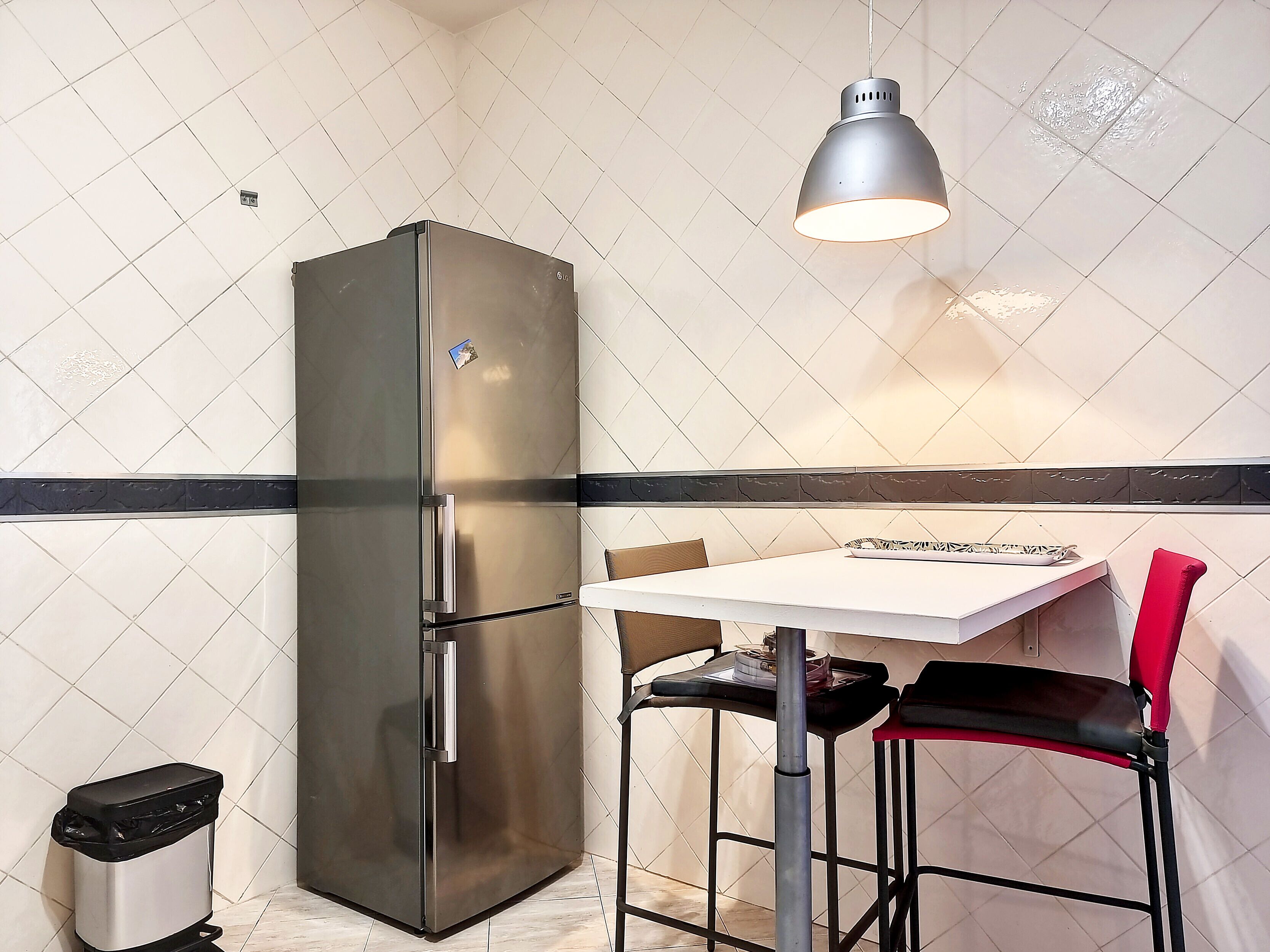 A compact kitchen with a stainless steel refrigerator, a white tiled backsplash, a small table with two chairs, and a trash bin.