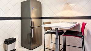 Een kleine keuken met een roestvrijstalen koelkast, een witte betegelde achterwand, een tafeltje met twee stoelen en een prullenbak.