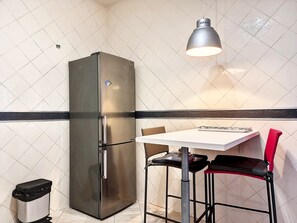A compact kitchen with a stainless steel refrigerator, a white tiled backsplash, a small table with two chairs, and a trash bin. - Le Mosala 2 - Climatisation - Vue mer (Saint-Leu)