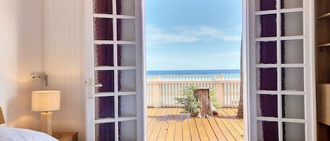 A room with a bed, a nightstand with a lamp, and a view of the beach through an open door.