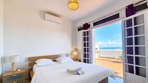 Een hotelkamer met een bed, nachtkastje, lamp en uitzicht op zee door een glazen deur.