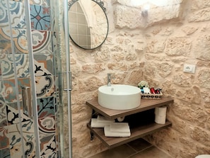 Une salle de bain avec un mur en pierre, un miroir rond, un lavabo blanc et une zone de douche carrelée.