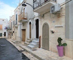 Una stretta via fiancheggiata da edifici tradizionali, con balconi dotati di ringhiere in metallo, una pianta in vaso e un cielo sereno.
