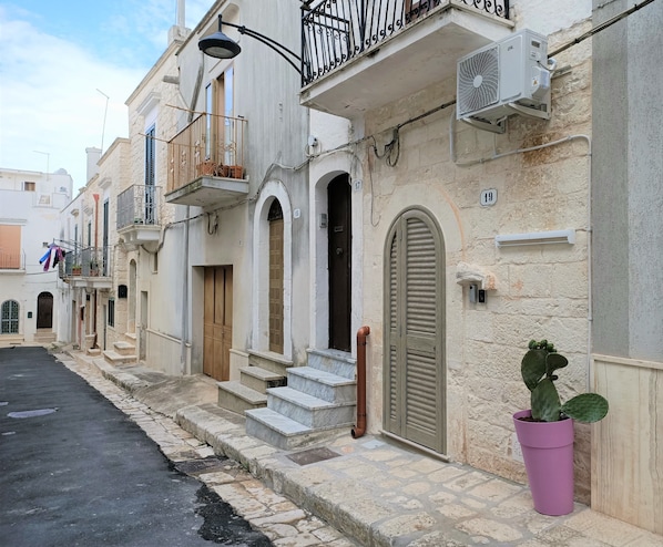 Une rue étroite bordée de bâtiments traditionnels, avec des balcons ornés de rampes en métal, un pot de fleurs et un ciel dégagé.