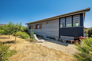A single-story house with a gravel driveway, a wooden deck, and a small garden area with shrubs and a tree. - Le Téno - Nature et détente - Wifi (Marzan)