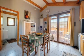 Un coin repas avec une table dressée pour quatre personnes, des chaises en bois, une horloge au mur et une porte-fenêtre coulissante donnant sur une terrasse extérieure.
