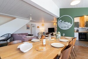 A dining area with a long wooden table set for a meal, a kitchen in the background, and a green wall with a circular sign. - Villa Insula - Proche plage - Jardin (Les Portes-en-Ré)