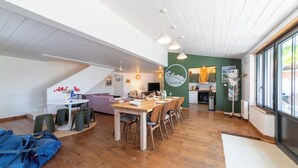 A spacious living area with a dining table, chairs, and a sofa. There is a kitchen area with a stove and cabinets. The room has a high ceiling with exposed beams and large windows. - Villa Insula - Proche plage - Jardin (Les Portes-en-Ré)