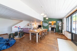 Ein großzügiger Wohnbereich mit Esstisch, Stühlen und einer Couch. Es gibt eine Küchenzeile mit Herd und Schränken. Der Raum verfügt über eine hohe Decke mit freiliegenden Balken und großen Fenstern.