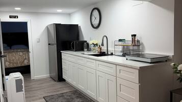 Uma cozinha compacta com armários brancos, uma geladeira preta, micro-ondas e uma pia com torneira. Há um relógio na parede e uma pequena planta no balcão.