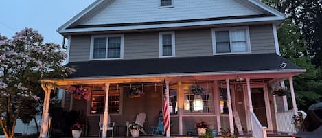A two-story house with a covered porch, a flag, and potted plants.