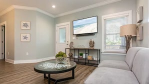 A living room with a flat-screen TV, a glass coffee table, a sofa, and a window with blinds.