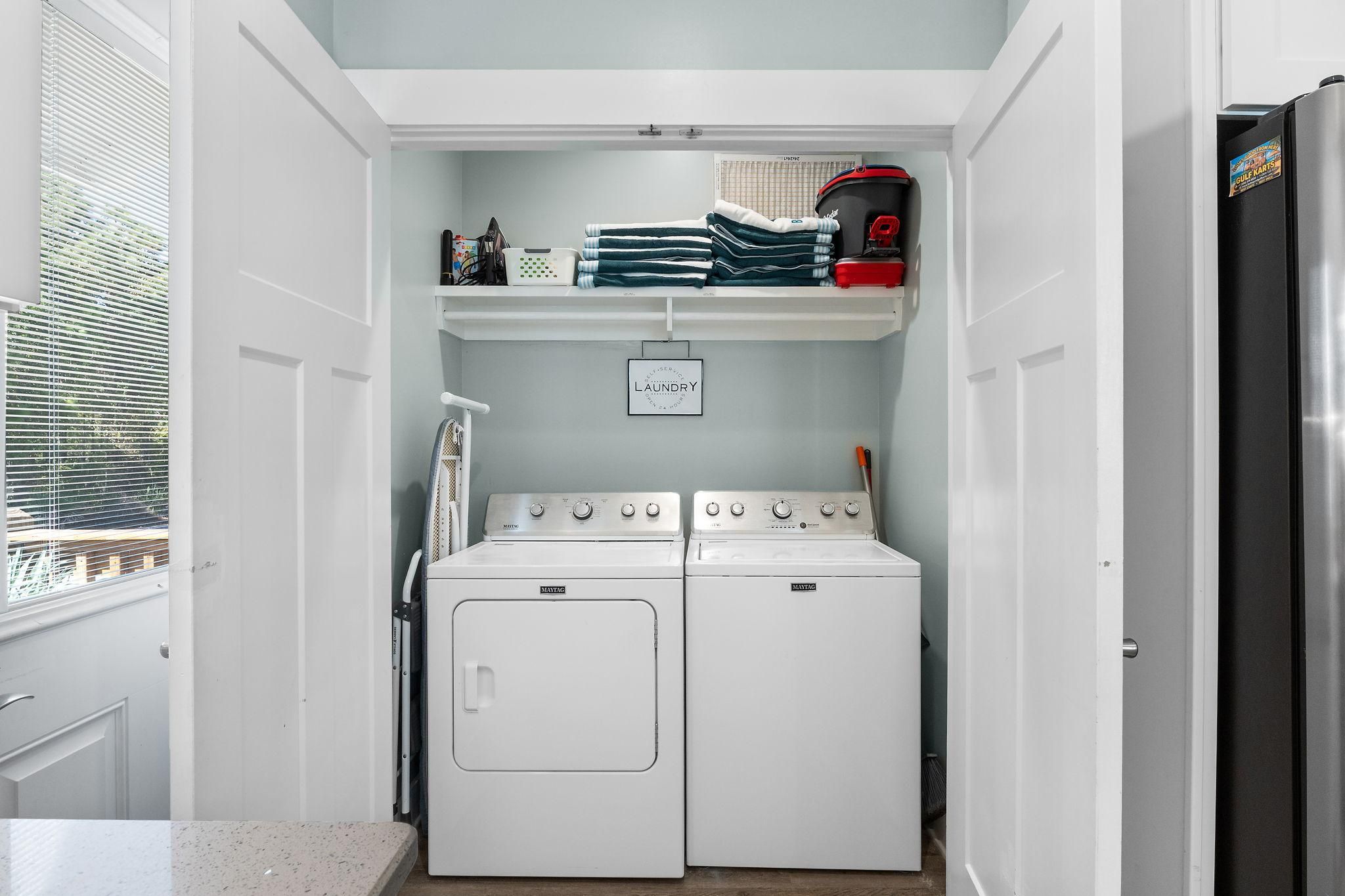 A laundry room with two white front-loading washing machines, a shelf with folded towels, and a sign indicating laundry services.
