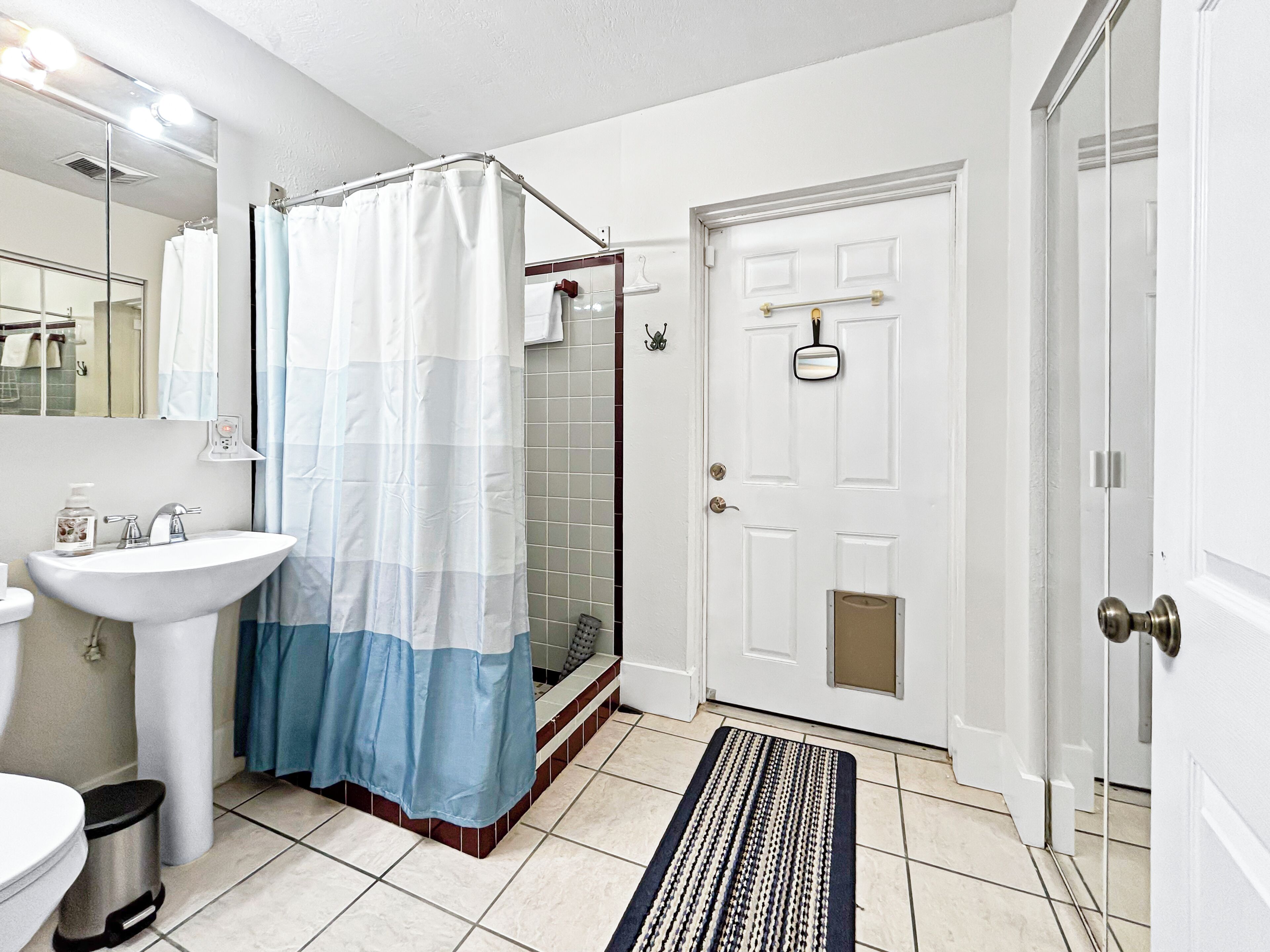Une salle de bain avec un lavabo blanc, une douche avec un rideau de douche bleu et blanc, et une porte blanche avec une petite fenêtre.