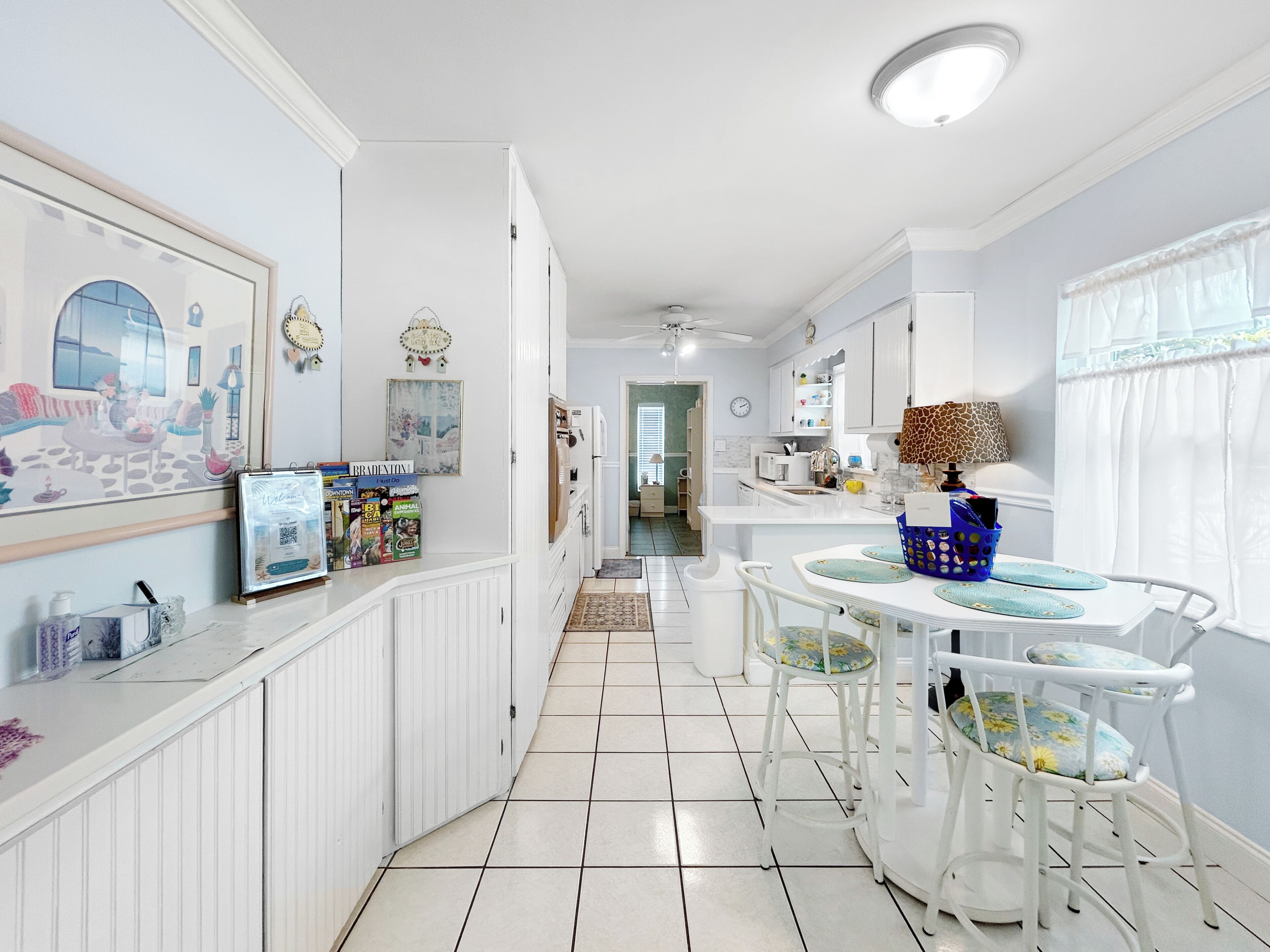A narrow hallway with a kitchen area on the right, featuring white cabinetry, a dining table with chairs, and a ceiling fan.