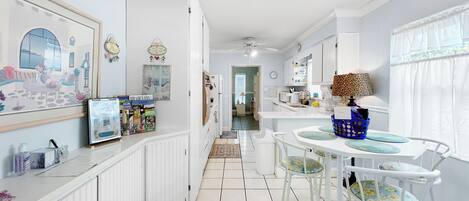 A narrow hallway with a kitchen area on the right, featuring white cabinetry, a dining table with chairs, and a ceiling fan.