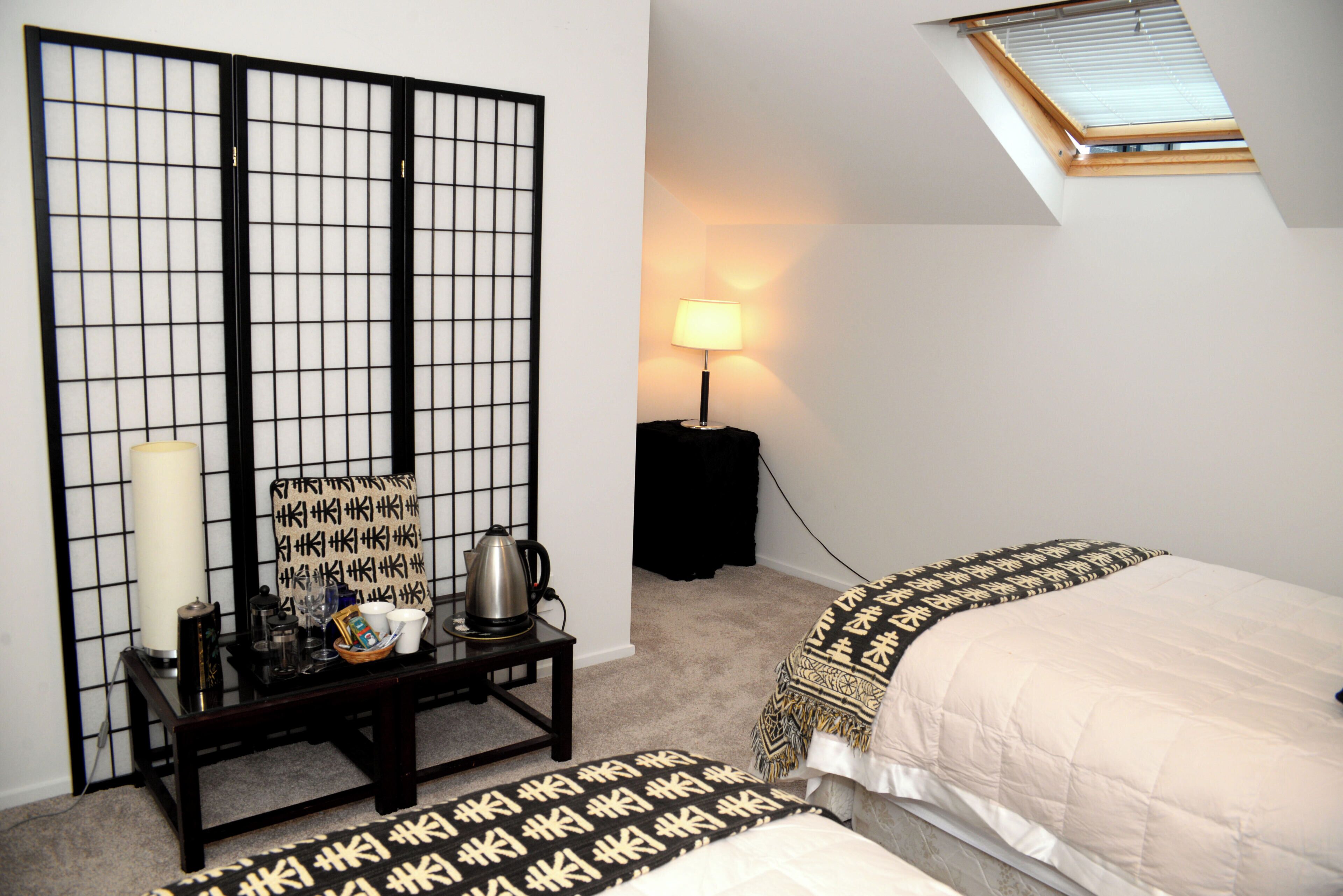 A bedroom with a bed, a nightstand with a lamp, a window with blinds, and a shoji screen.
