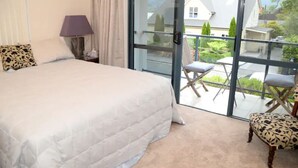 A bedroom with a large bed, a nightstand, a lamp, and a view through a glass door to an outdoor seating area. - Spacious Villa in the Heart of Akaroa (Akaroa)