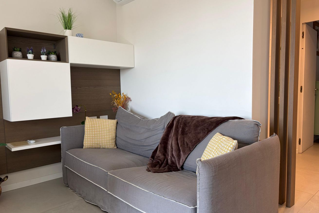 Un salon moderne avec un canapé gris, une étagère blanche fixée au mur, agrémentée de plantes, et une porte située à droite.